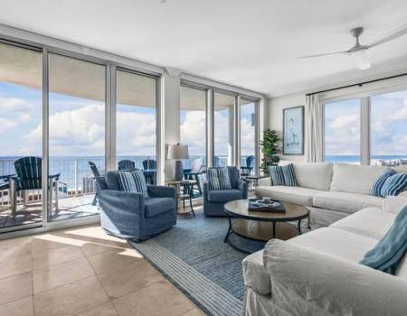 Living area with a white couch, a blue rug, a round coffee table and two swivel chairs looking out at the water with a balcony